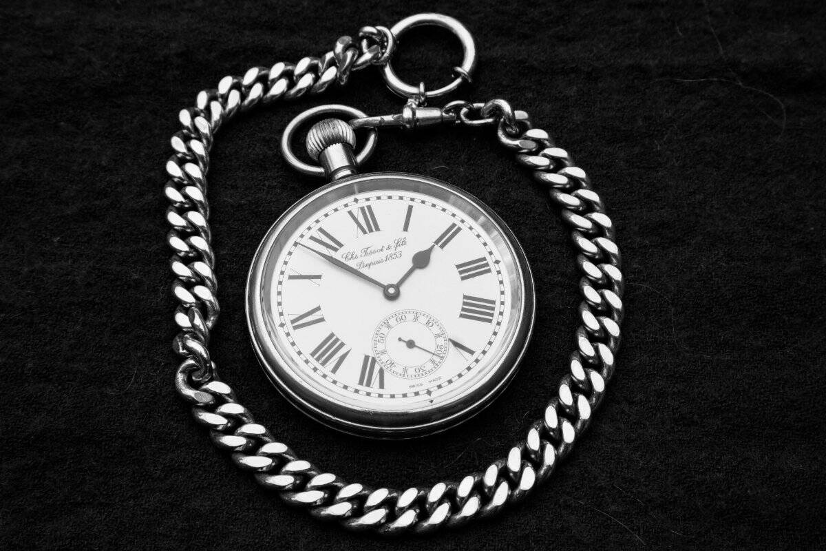 Reloj de bolsillo con números romanos y una cadena gruesa sobre fondo negro